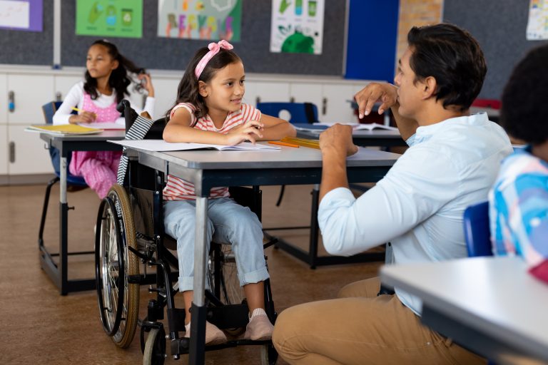 Happy biracial male teacher with girl in wheelchair in class at elementary school. School, learning, childhood, disability, inclusivity, teaching and education, unaltered