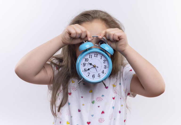 looking at camera little school girl wearing white t-shirt covered face with alarm clock on isolated white background