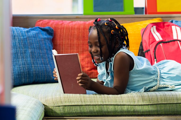 Happy african american schoolgirl reading book in colourful room at elementary school. Education, childhood, development, learning and school, unaltered